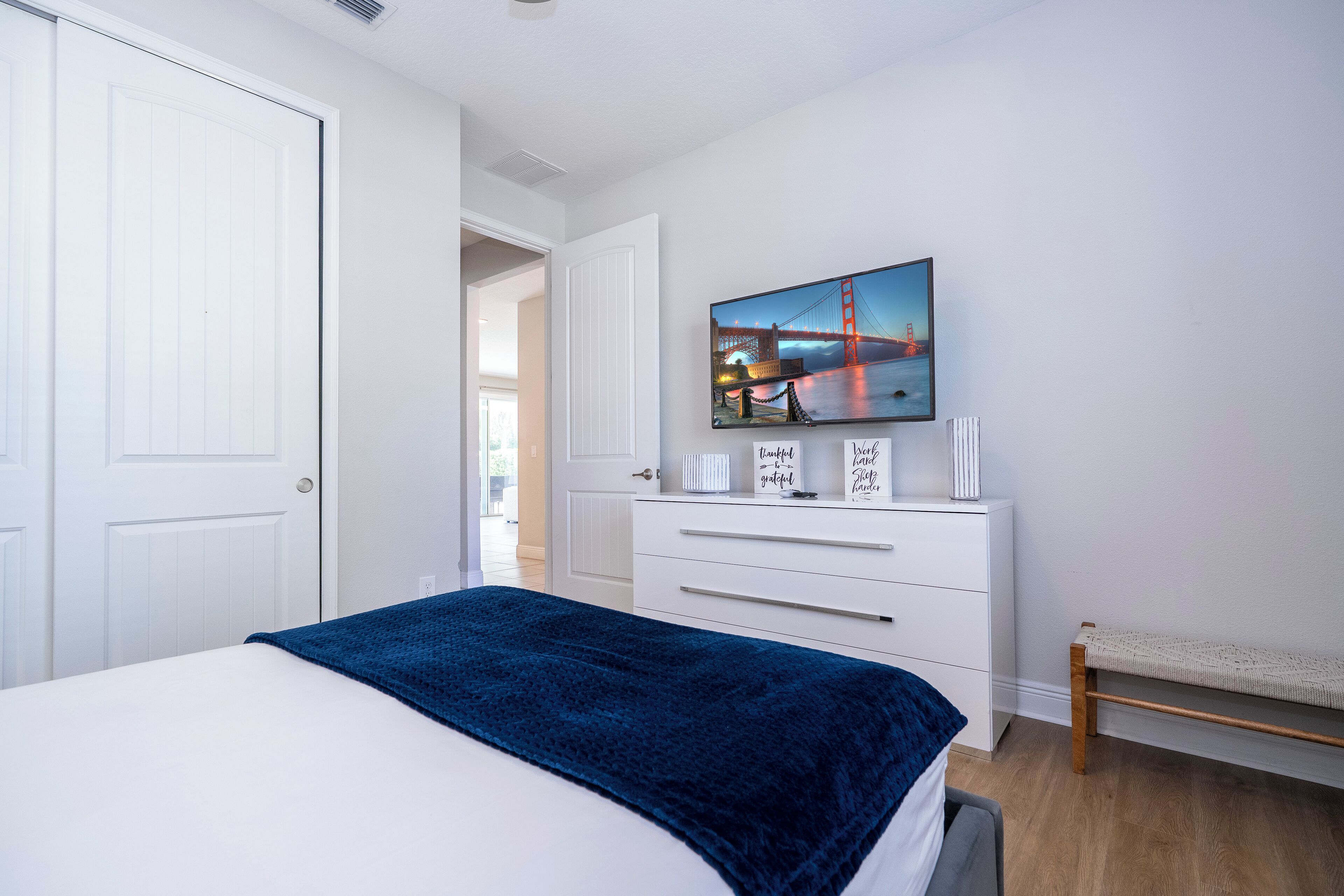 A bedroom with a bed, a dresser, a bench, and a painting of a bridge.