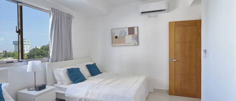 A modern hotel room with a large window, a bed with white bedding and blue pillows, a bedside table with a lamp, and an air conditioning unit on the wall.