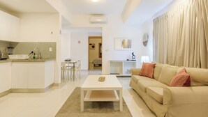 Un salon moderne avec une table basse blanche, un canapé beige et un coin cuisine doté de meubles blancs et d’un plan de travail.