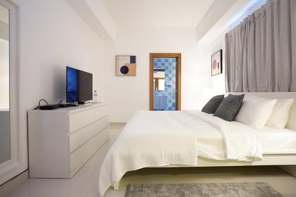 Un dormitorio moderno con una cama, un televisor sobre un aparador, y un baño visible a través de una puerta abierta.