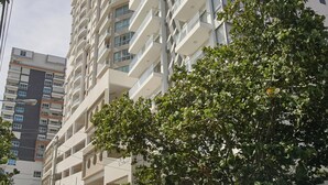 Un immeuble résidentiel à plusieurs étages doté de balcons, avec un arbre au premier plan et une rue où est garée une voiture.