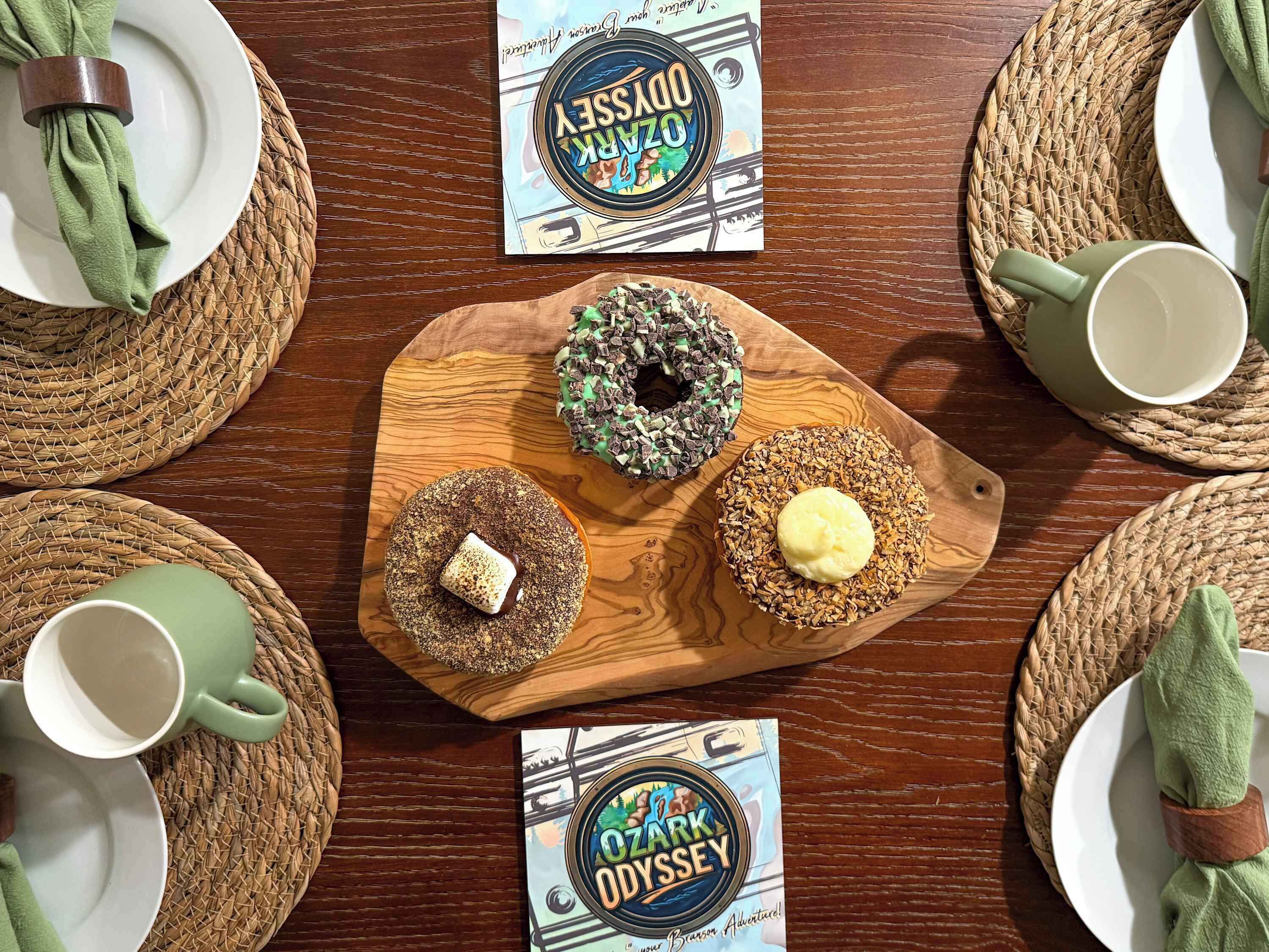 Une planche en bois sur laquelle sont disposés trois beignets, une serviette verte et deux sets de table, le tout posé sur une table en bois.