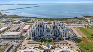 A modern hotel with a central courtyard, multiple floors, and a view of the sea and a bridge in the background. - 2-Level Beachfront Resort Condo w/Views of Beach & Bay! (South Padre Island)