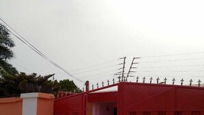 Une entrée fermée par un portail rouge, un chemin en béton et un bâtiment à la façade rose, visible à travers le portail.