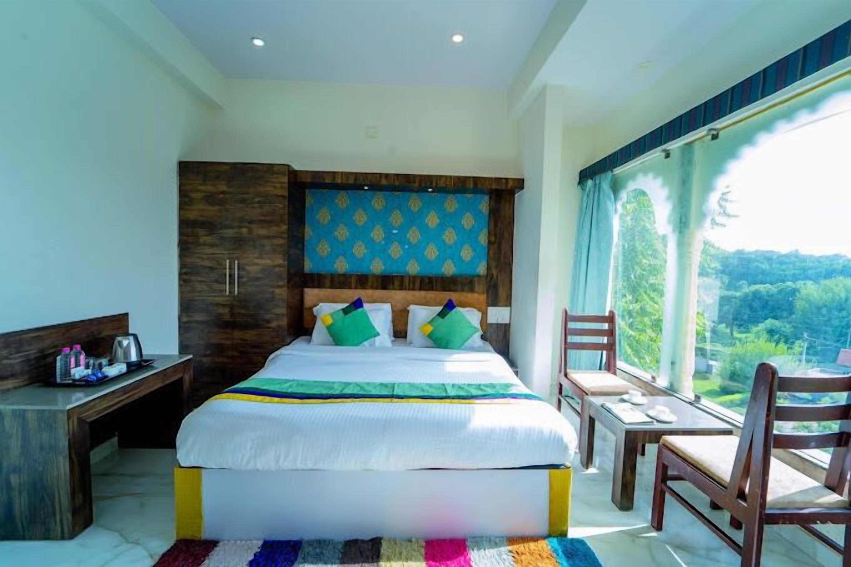A hotel room with a large bed, a wooden wardrobe, a small table with chairs, and a view of greenery through the window.