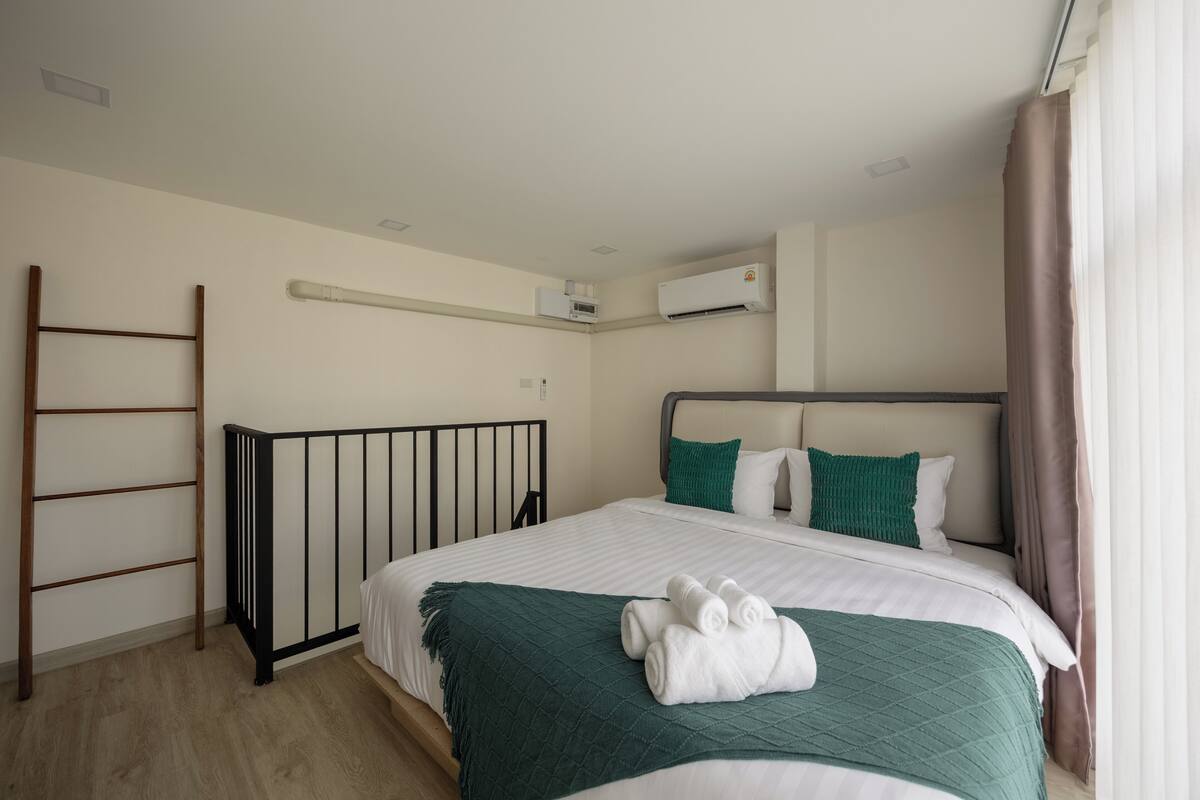 Cama bem arrumada com manta verde e toalhas dobradas, berço de metal preto e unidade de ar condicionado branca na parede.