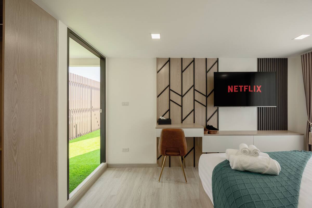 Quarto de hotel moderno com uma grande tela de TV exibindo "NETFLIX", uma escrivaninha, uma cadeira e uma cama com um edredom verde.