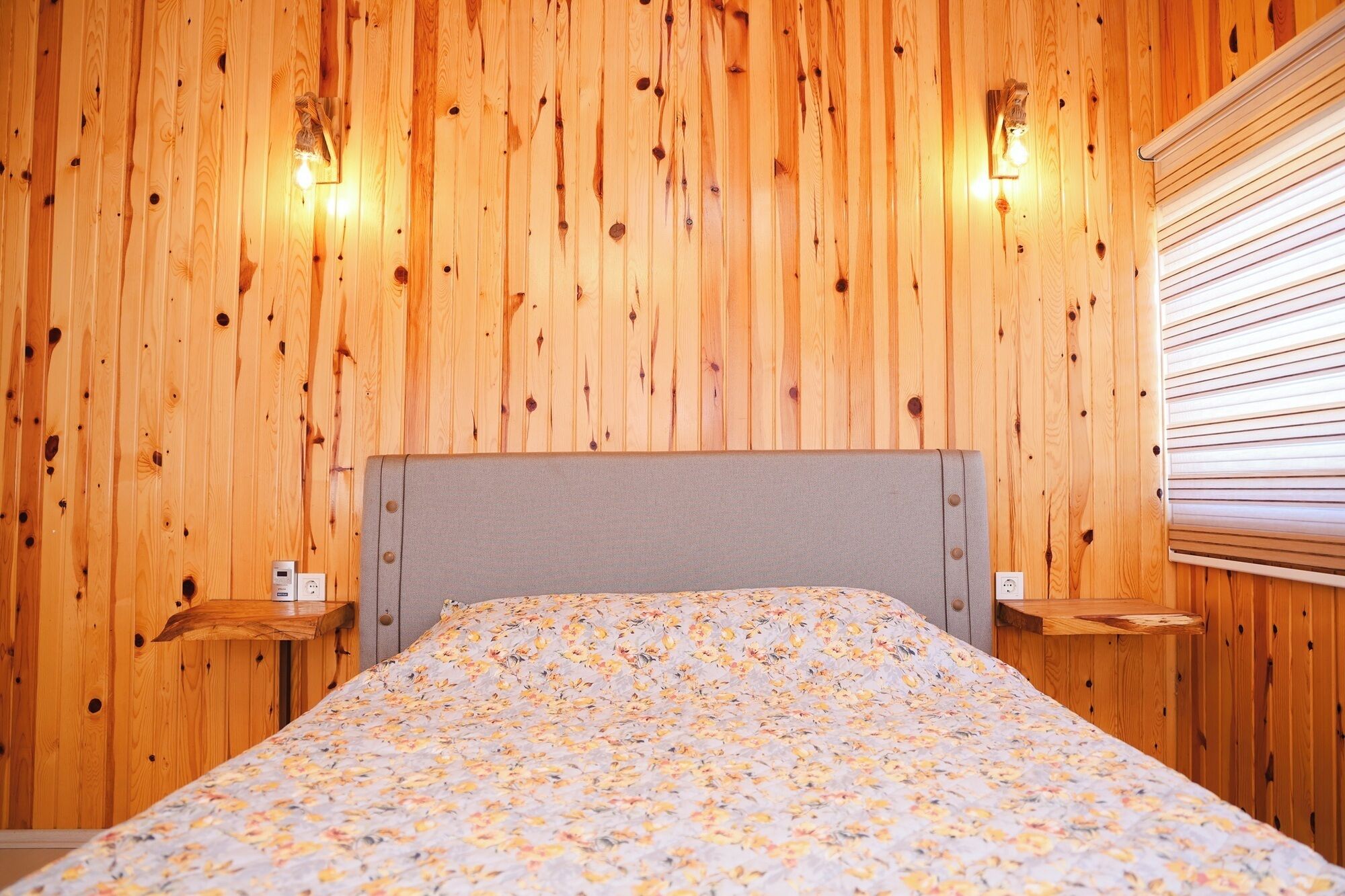 Une chambre avec un mur lambrissé en bois, un lit recouvert d’une couvre-lit à motifs floraux, deux tables de chevet et une fenêtre avec des stores.