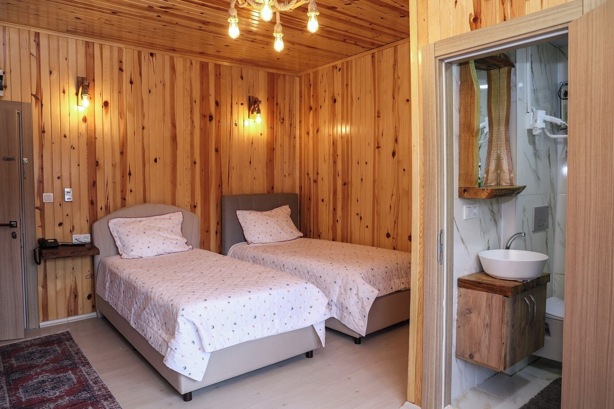 A wooden-paneled room with two beds, a bedside table with a phone, and a bathroom with a sink and mirror.