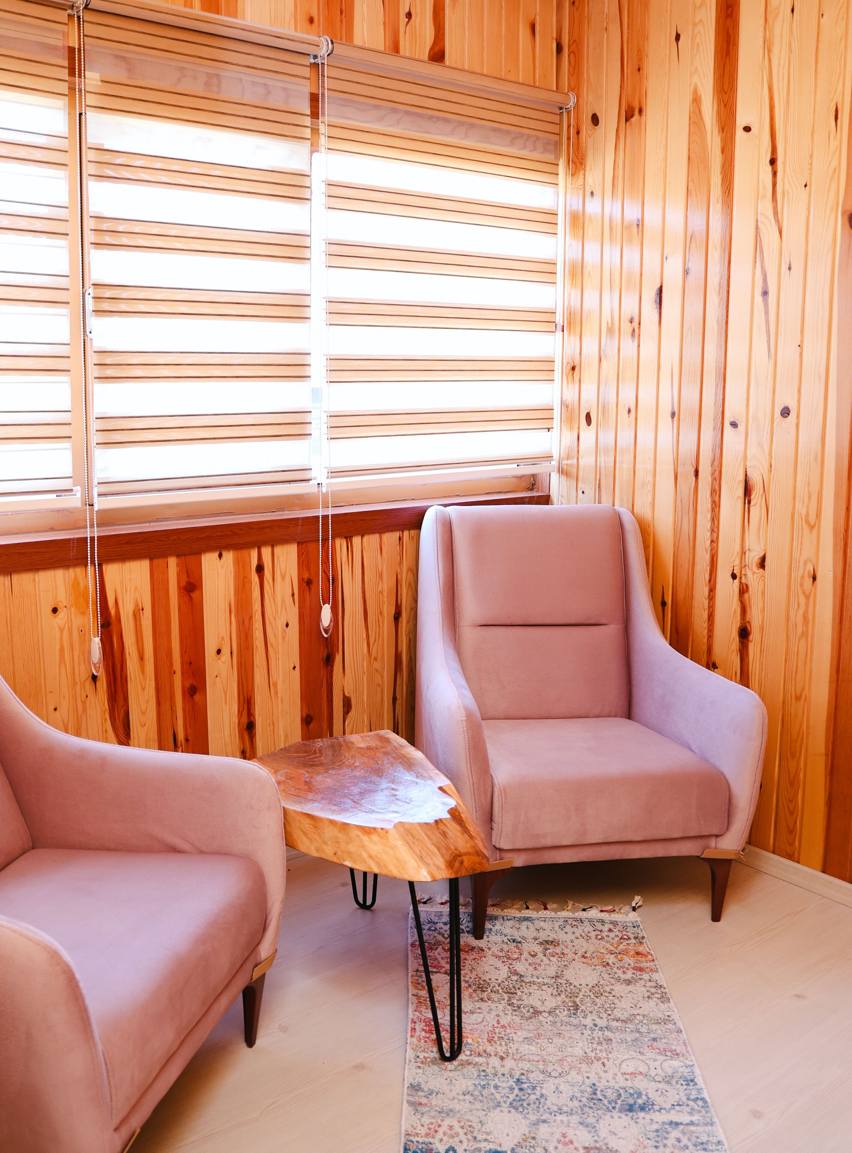 Une pièce chaleureuse aux murs en bois, avec deux fauteuils roses, une petite table en bois et une grande fenêtre avec des stores.