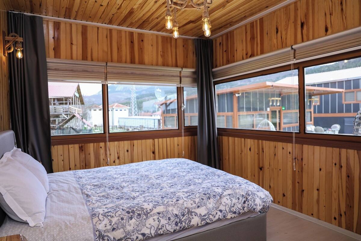 Un dormitorio con cama, paredes de madera y ventanas amplias con vistas a edificios y montañas.