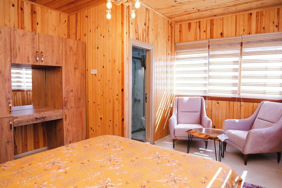 Una habitación de madera con una cama, dos sillas, una mesita y una zona de ducha.