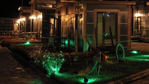 A modern building with a glass door entrance, outdoor seating, and a small garden area with green lighting. - Horma Kanyon Otel (Pinarbasi)