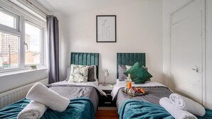 A hotel room with two beds, each with a headboard, a window with curtains, a framed picture on the wall, and a tray with fruits and a bottle on one bed. - Hillside Haven in Manchester (Manchester)