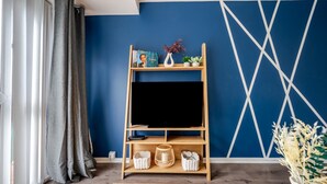 A living room with a TV on a wooden stand, a blue wall with geometric patterns, a window with curtains, and a wooden coffee table. - Hillside Haven in Manchester (Manchester)