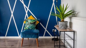 A room with a blue geometric wall design, a blue velvet chair with yellow and green pillows, a potted plant on a white radiator, and a black metal side table. - Hillside Haven in Manchester (Manchester)