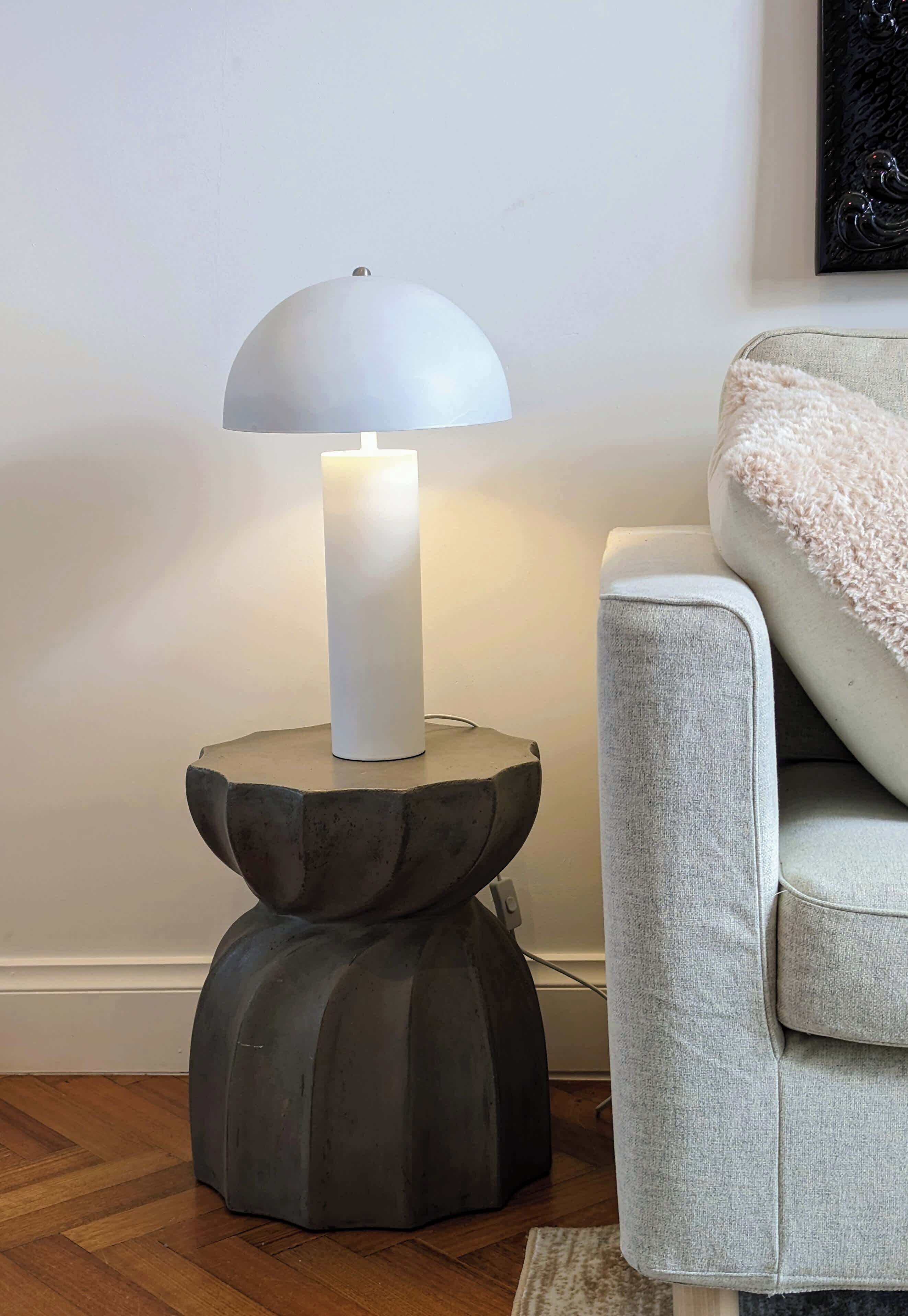 Eine moderne Stehlampe mit weißem Lampenschirm und dunklem Fuß auf einem runden Sockel, neben einem hellen Sofa mit weicher Decke.
