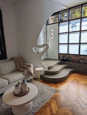 A modern living room with a curved staircase, a sofa, a coffee table, and a large window with a view of trees. - European Style Escape in Geelong (Geelong)