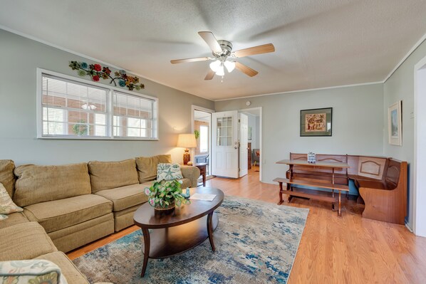 A living room with a sofa, coffee table, and wooden bench. There is a ceiling fan, a window with blinds, and a door leading to another room.