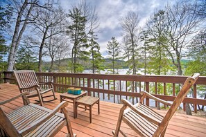 A wooden deck with seating and a table, overlooking a lake with trees and hills in the background.