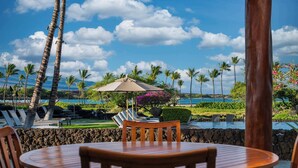 A wooden table and chairs in a tropical resort setting with a pool, palm trees, and a clear sky. - Luxurious Kolea Kai Villa, Private Lanai w/ Ocean Views, Beach Club, Kona Coffee (Waikoloa Village)