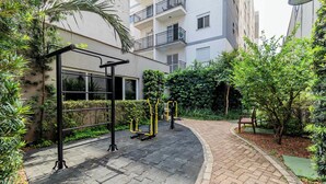 An outdoor gym area with exercise equipment, surrounded by greenery and a paved walkway.