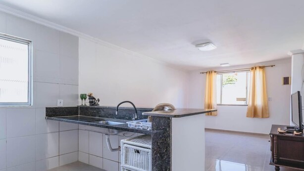 A compact kitchen with a black countertop, a sink, and a small island. There is a window with curtains and a television on a wooden stand. - Apartamento no Rio próximo à Lagoa da Tijuca (Rio de Janeiro)