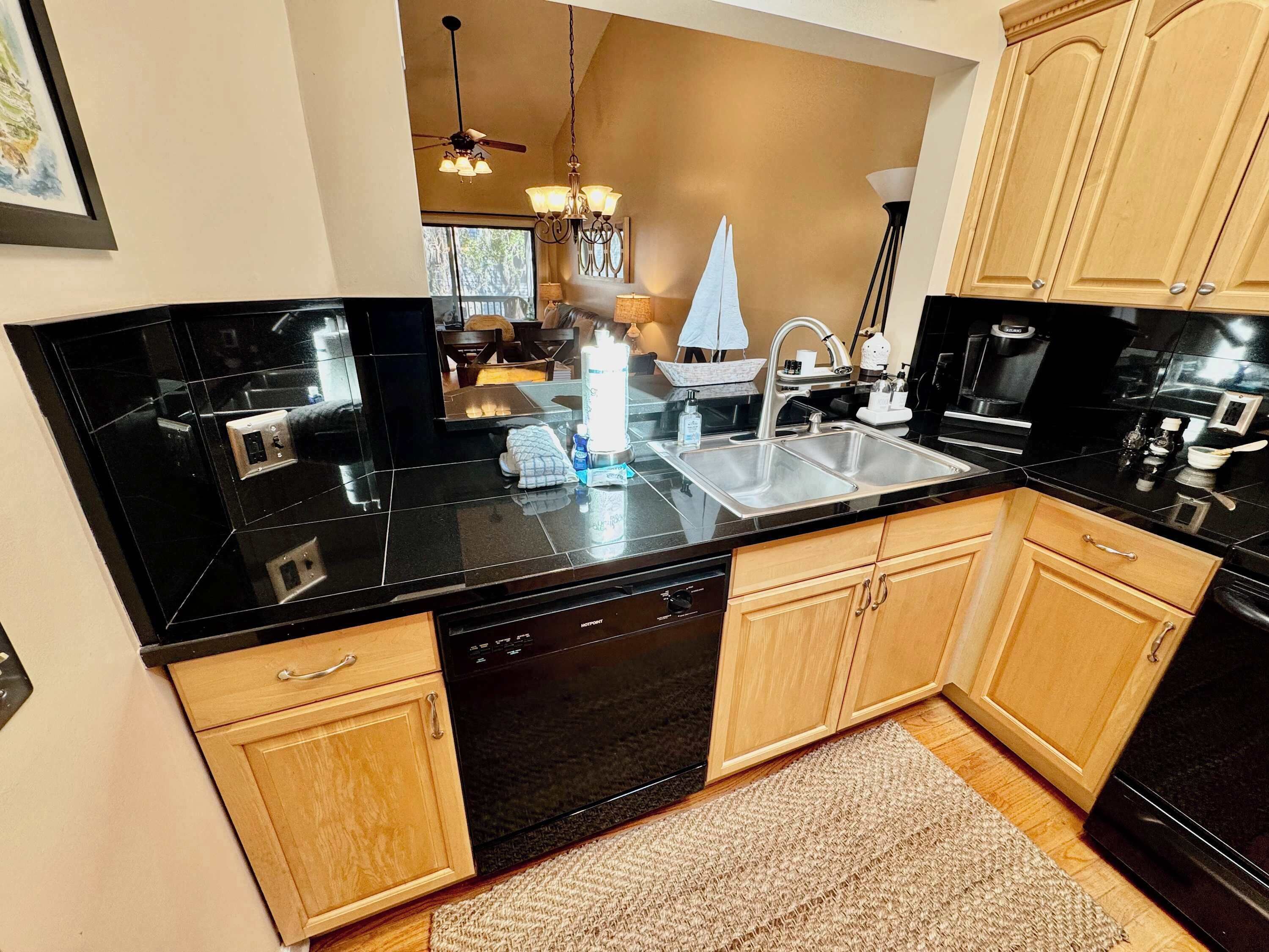 A modern kitchen with black countertops, wooden cabinets, a dishwasher, and a large window.