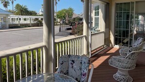 A covered outdoor area with a view of a parking lot and palm trees. There are white wicker chairs and a glass table.