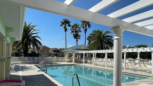 A pool area with lounge chairs and a covered section, surrounded by palm trees and buildings.