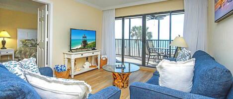 A living room with blue sofas, a glass coffee table, and a TV displaying a beach scene.
