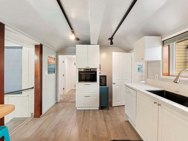 A compact kitchen with white cabinets, a built-in oven, and a microwave. There is a wooden dining table and a blue stool in the foreground.