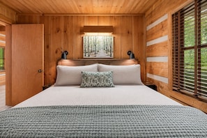 A wooden cabin bedroom with a large bed, two bedside lamps, a forest painting on the wall, and a window with blinds. - Summer Sale! Creek Views-Hot Tub-Games-Fire Pit (Pigeon Forge)