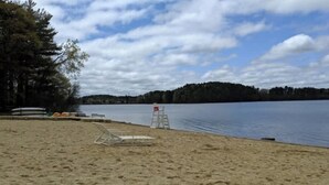 A sandy beach with a lifeguard chair and a small structure, likely a lifeguard station, near a calm lake with a forested hill in the background. - Spacious 4 bedroom home with large yard (Wayland)