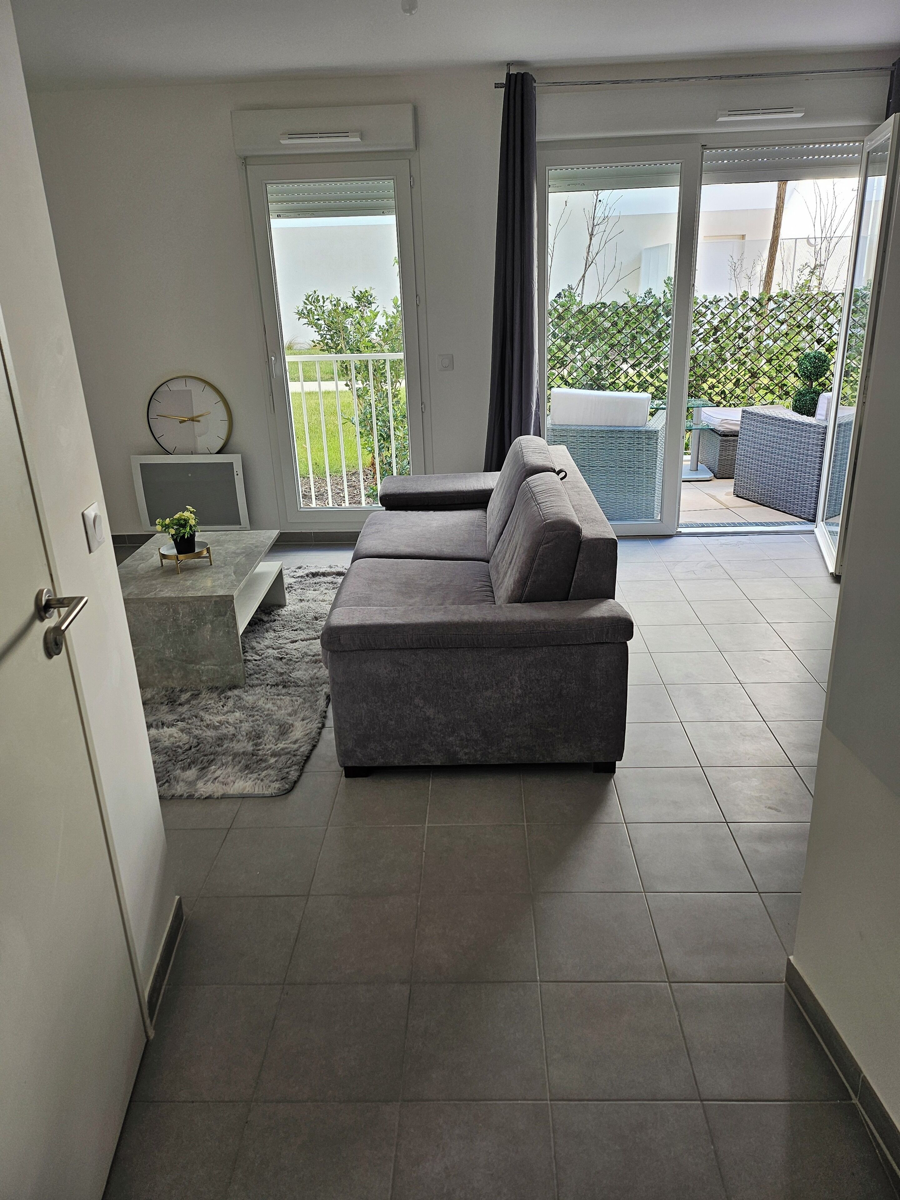 A modern living room with a grey sectional sofa, a small white side table with a plant, and a grey rug. There is a sliding glass door leading to an outdoor patio area with a table and chairs.