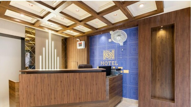 A hotel reception area with a wooden reception desk, a blue wall with the hotel logo, and a ceiling with wooden beams and recessed lighting. - Hotel Noble Inn (Ahmedabad)