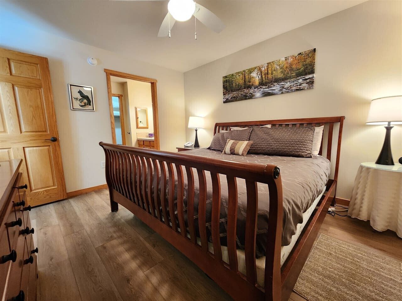 A bedroom with a wooden bed, a nightstand, a lamp, a painting, and a door leading to another room.
