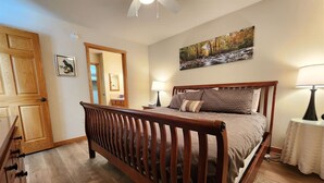 Un dormitorio con una cama de madera, una mesita de noche, una lámpara, un cuadro y una puerta que da a otra habitación.