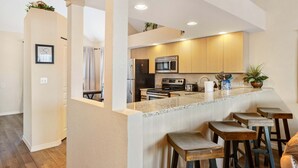 Eine moderne Küche mit Holdekor-Schränken, einem Edelstahlkühlschrank und einer eingebauten Mikrowelle. Es gibt Barhocker und eine Arbeitsplatte mit Spüle.