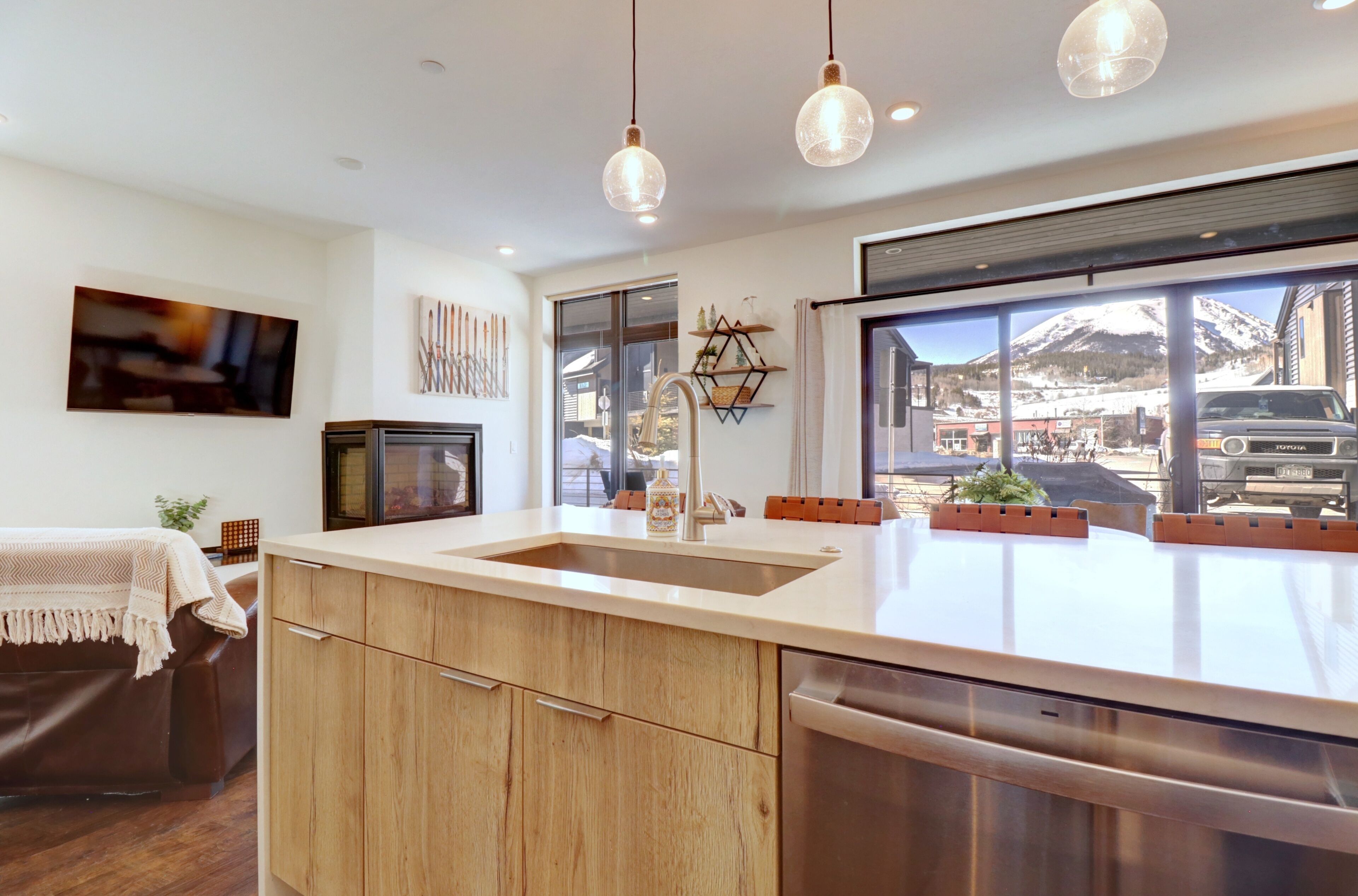 A modern kitchen with a large island, wooden cabinets, and a stainless steel dishwasher. There is a large window offering a view of a snowy mountain and a parked car outside.