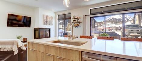 A modern kitchen with a large island, wooden cabinets, and a stainless steel dishwasher. There is a large window offering a view of a snowy mountain and a parked car outside.