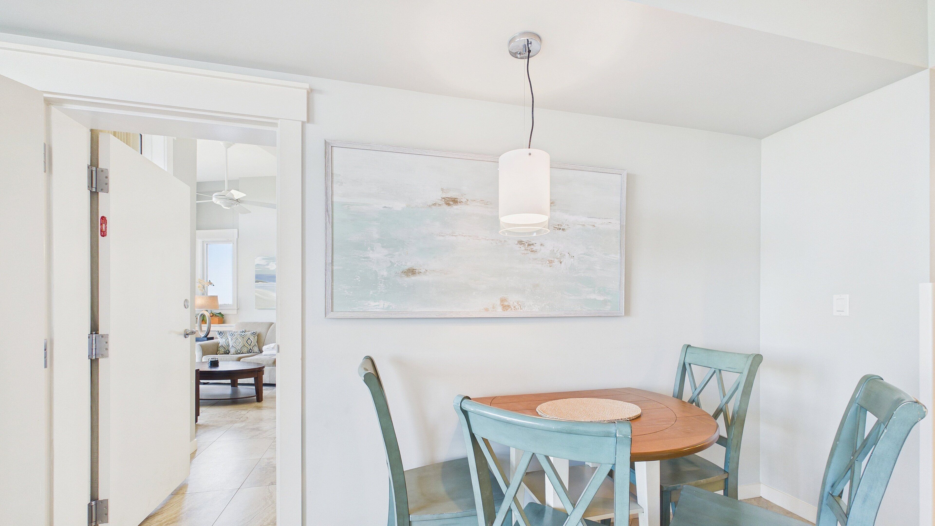 A dining area with a round wooden table, four blue chairs, a hanging pendant light, and a large abstract painting on the wall.