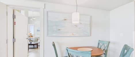 A dining area with a round wooden table, four blue chairs, a hanging pendant light, and a large abstract painting on the wall.