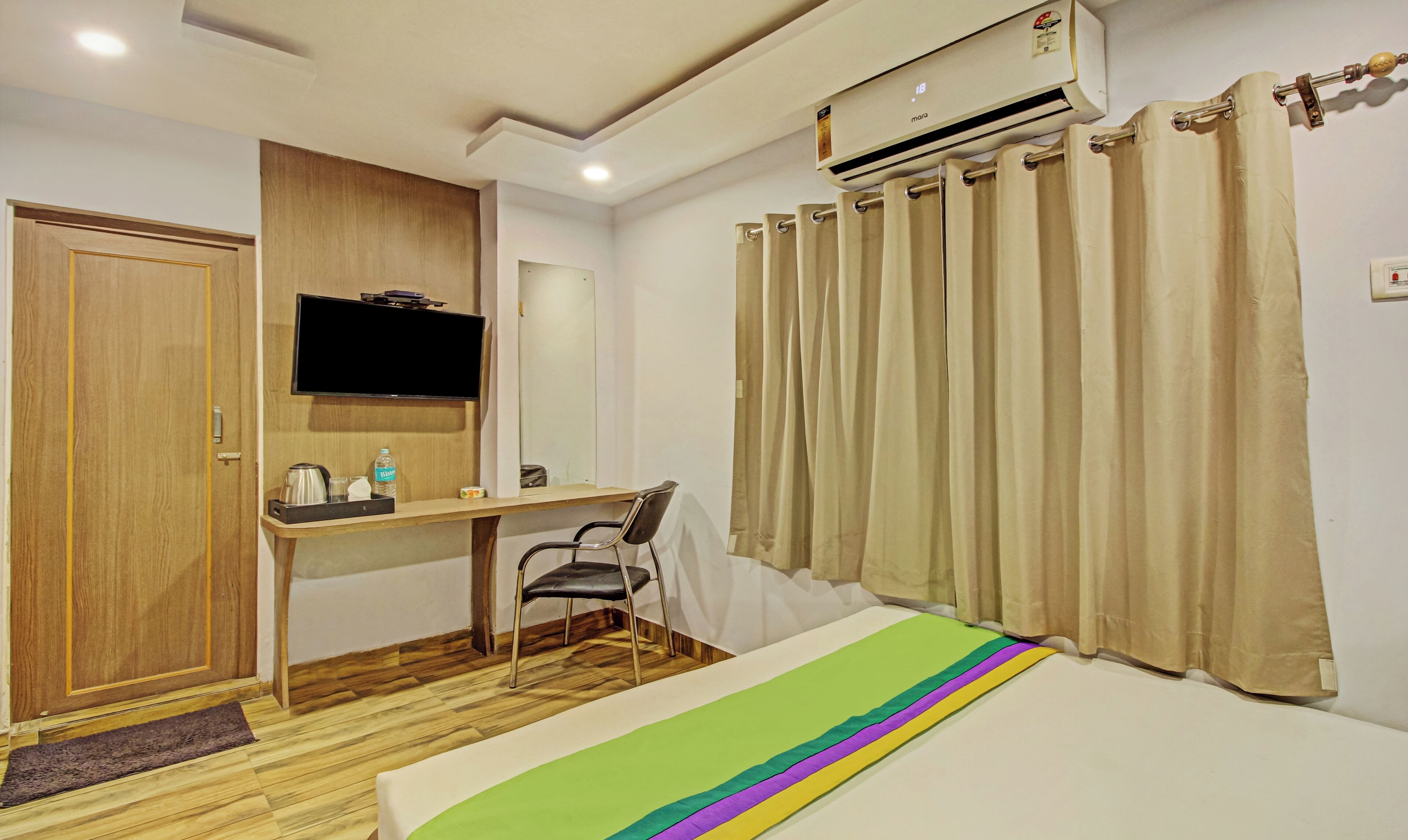 A hotel room with a wooden desk, a television, a bed with a colorful blanket, an air conditioning unit, and beige curtains.