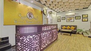 Un área de recepción con un letrero amarillo que dice "Hotel Royal Crowne", un mostrador de recepción, un sofá con cojines amarillos y obras de arte enmarcadas en las paredes.