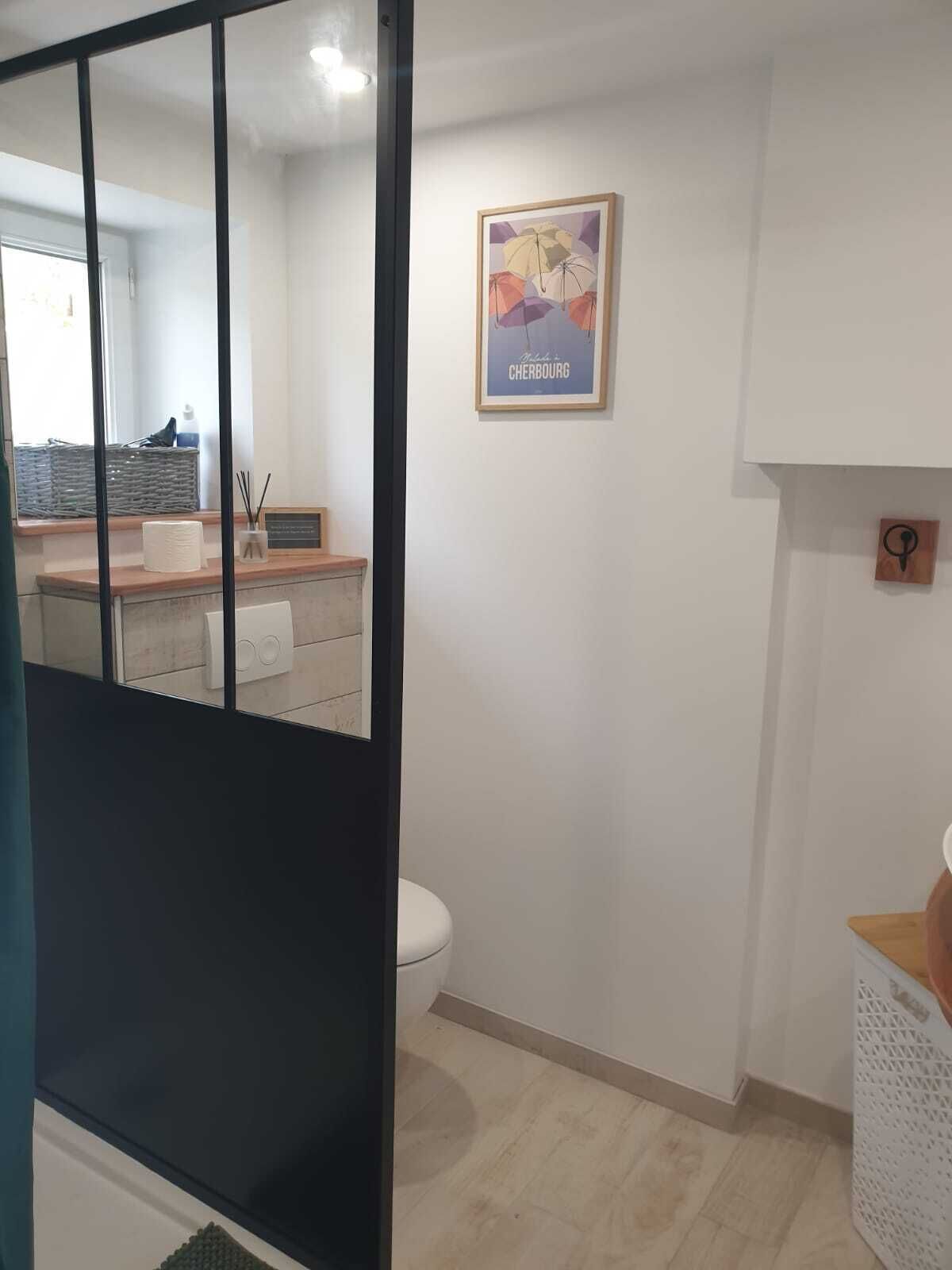 A modern bathroom with a toilet, a sink, and a shower area. There is a framed poster on the wall and a small wooden shelf.