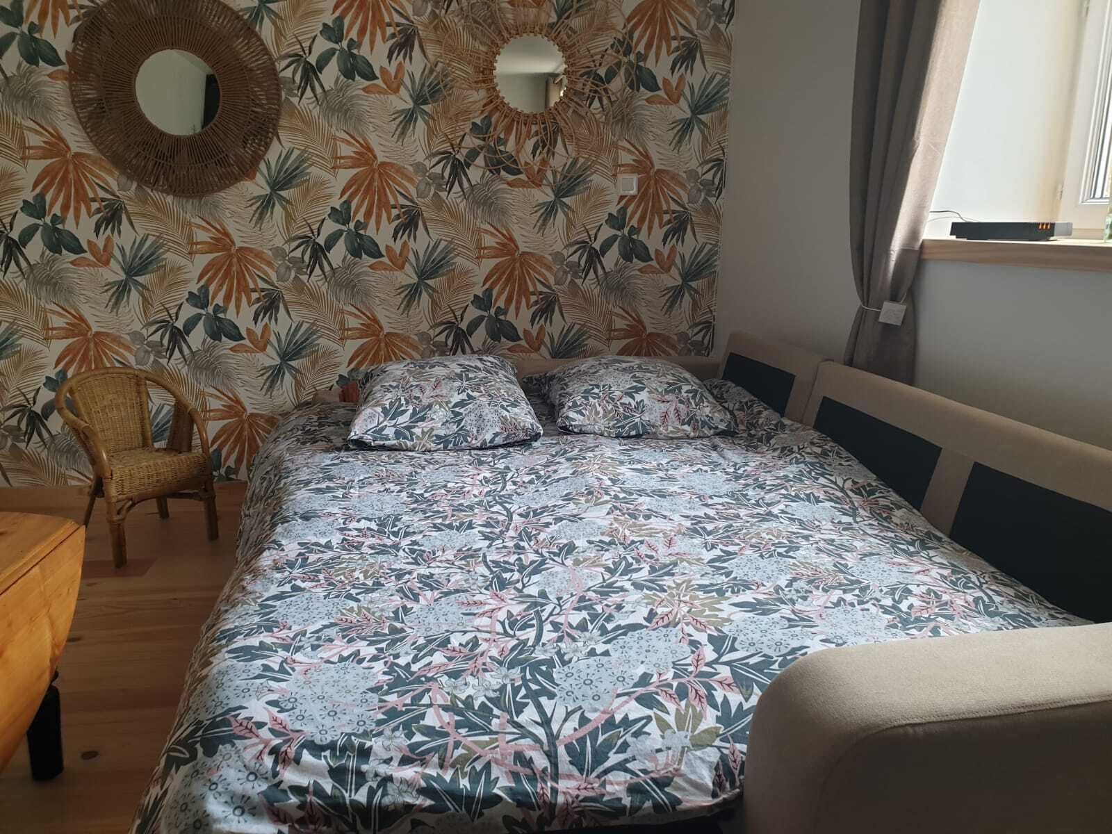 A bedroom with a bed, two pillows, a wicker chair, a round mirror, and a window with curtains.