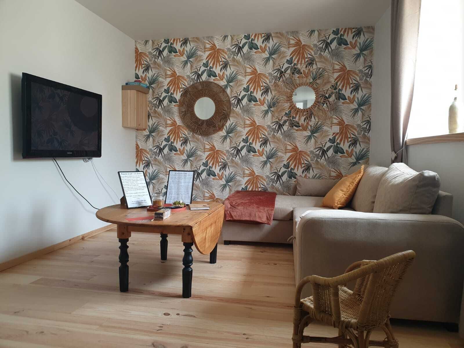 Una sala de estar con piso de madera, un espejo redondo, un sofá, un sillón de mimbre y una pared con estampado de hojas tropicales.