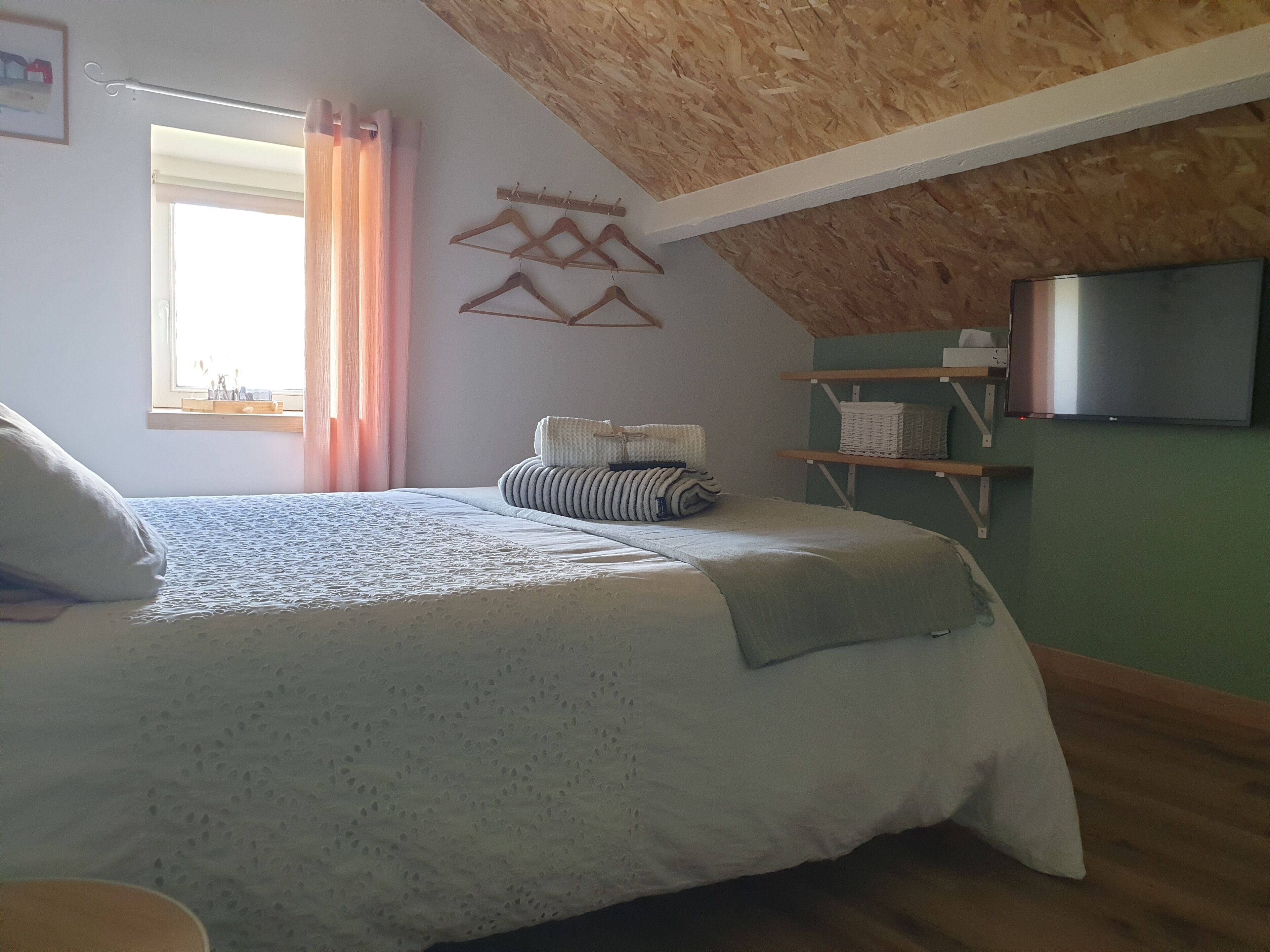 Een slaapkamer met een schuine hemel, een bed met een beddegoed met patroon, een raam met roze gordijnen en een wandtelevisie.