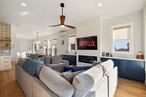 Ein modernes Wohnzimmer mit einem grauen Ecksofa, einem Deckenventilator, einem an der Wand montierten Fernseher und einer Küchenzeile im Hintergrund.